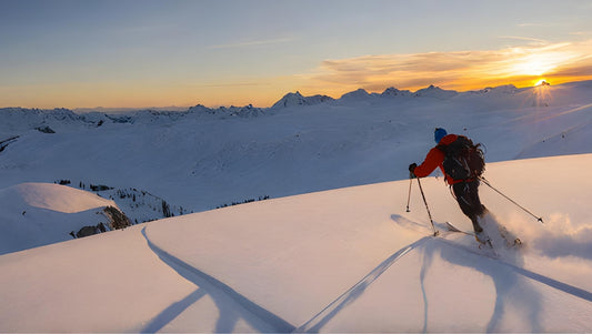 A skier gracefully descends a mountain slope as the sun sets in the background, casting a warm glow over the snowy landscape.
