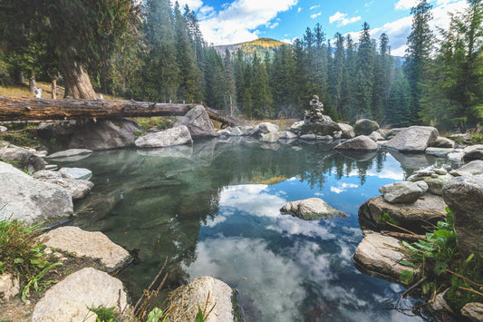 Serene mountain hot spring surrounded by rocks and trees, reflecting the sky.