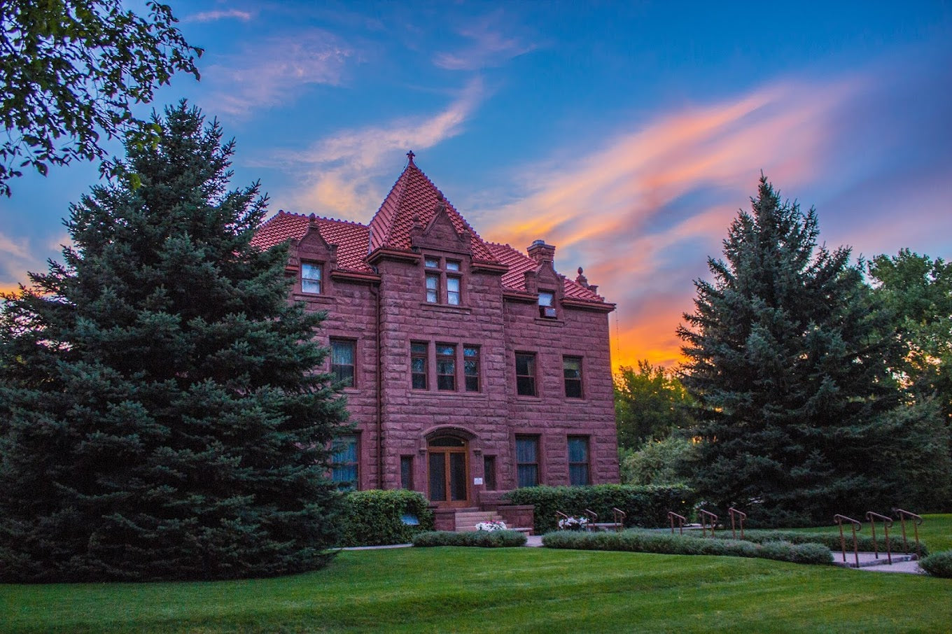 A sunset casts a warm glow on a large brick house featuring a striking red roof.