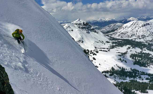 A skier descends a steep, snowy mountain slope with scenic peaks in the background.