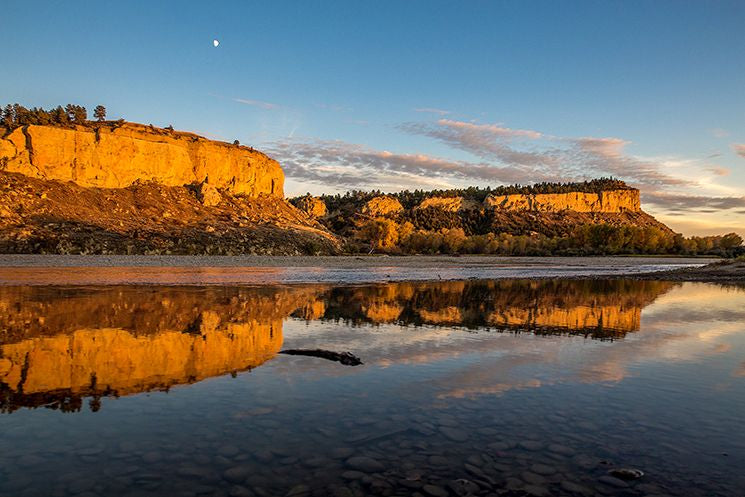 Golden cliffs reflect in a calm river at sunset, with a clear sky and a crescent moon overhead.