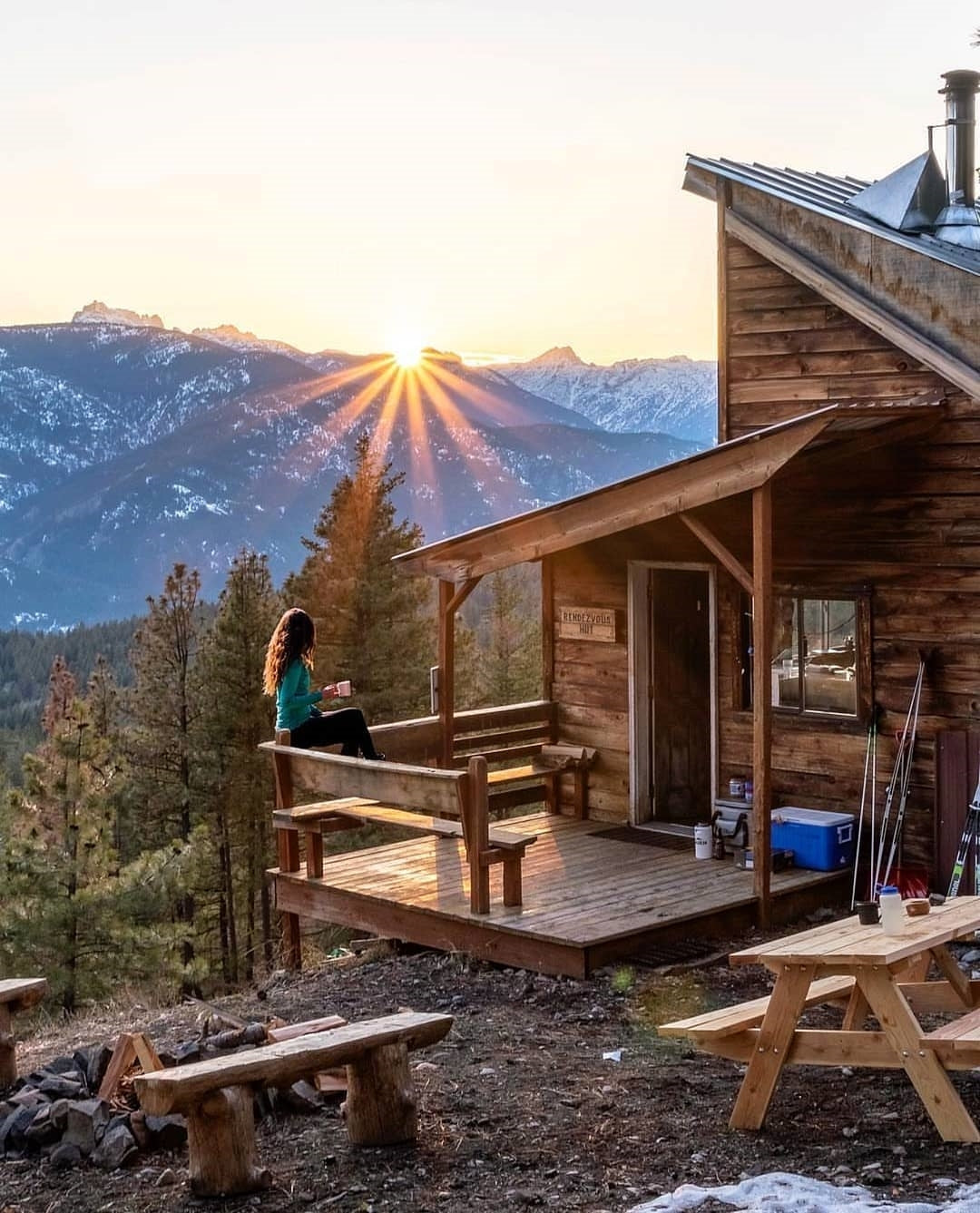 A woman sits on the porch of a rustic wooden cabin, enjoying a beverage as the sun sets over the snow-capped mountains in the background, creating a serene and picturesque scene.