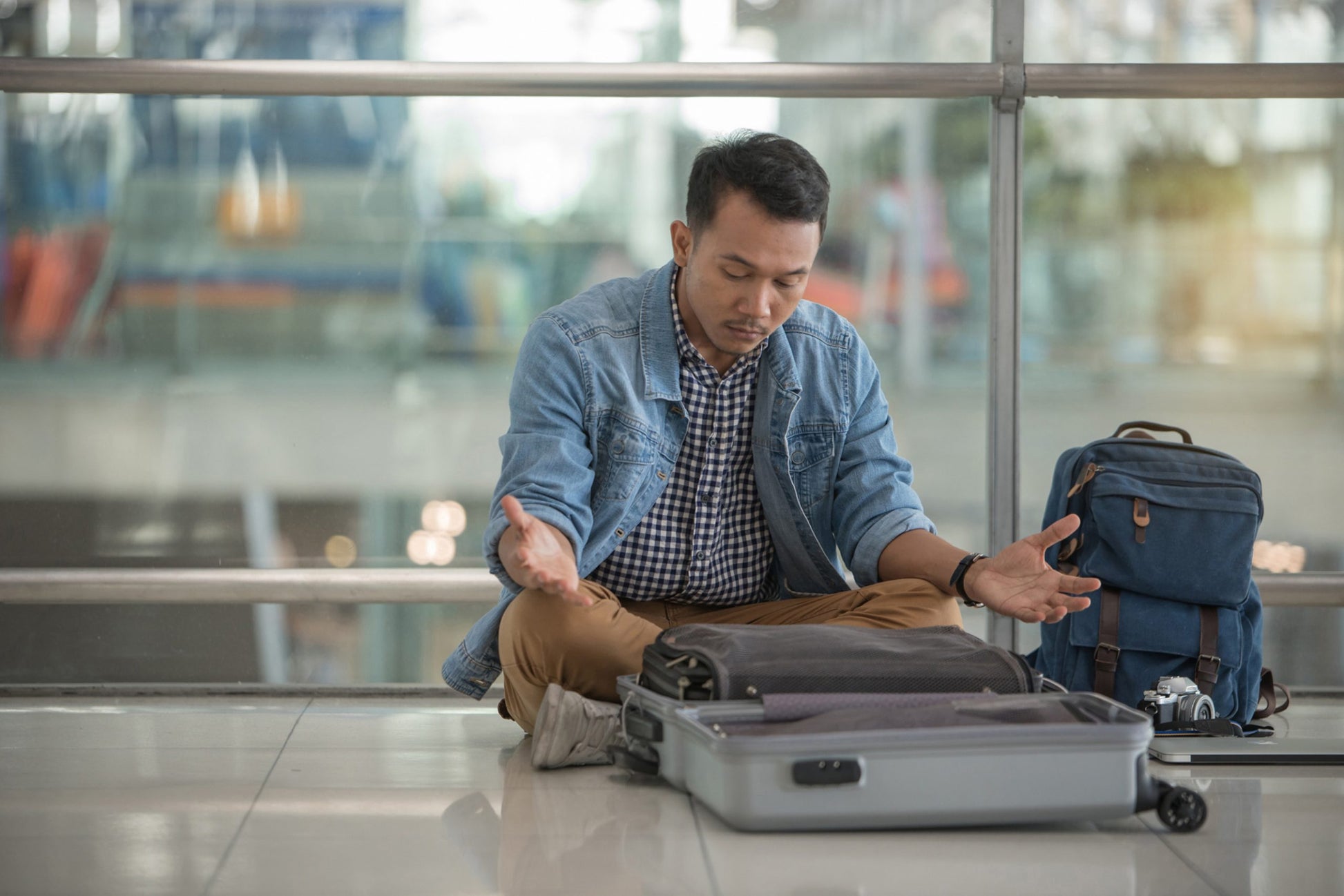 A frustrated man sits on the floor of an airport with his open suitcase in front of him, appearing concerned about his travel situation. He has a denim jacket, khaki pants, and a blue backpack beside him.