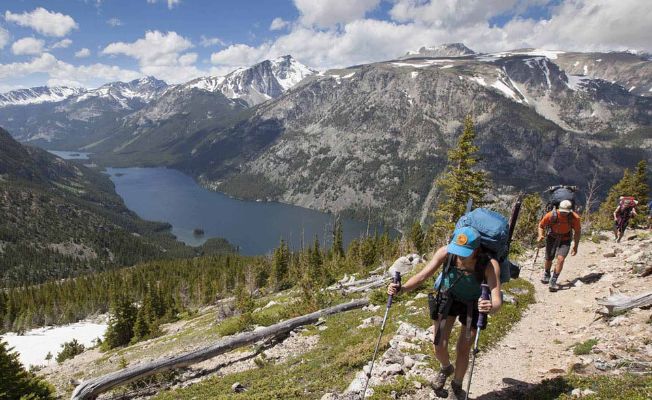 Hikers ascending a mountain trail with a scenic lake and snow-capped peaks in the background.