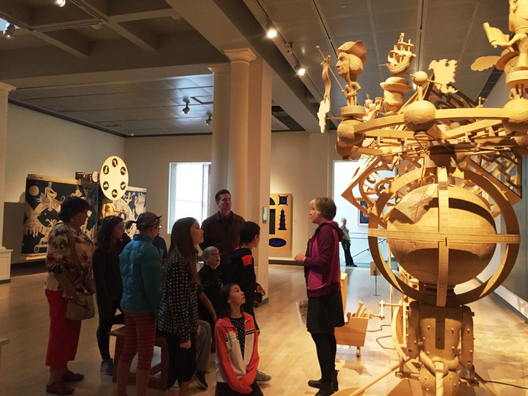 A group of people listens to a guide in an art gallery, admiring a large wooden sculpture.
