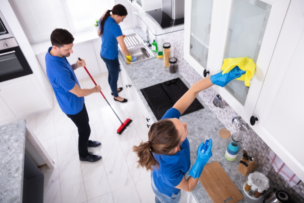 Two professional cleaners in blue uniforms actively cleaning a bright kitchen, one mopping the floor and the other wiping the cabinet glass doors, depicting a thorough and efficient cleaning service in a residential setting.