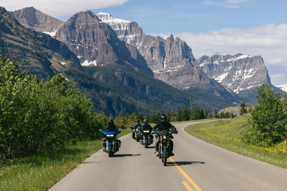 Group of motorcyclists riding on a scenic mountain road with towering peaks in the background.