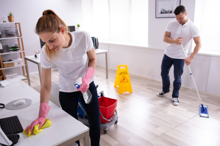  A professional cleaning team working in an office environment, with a woman wiping down a desk while wearing pink gloves and a man mopping the floor in the background, emphasizing a clean and well-maintained workspace with a "Wet Floor" caution sign visible to ensure safety.