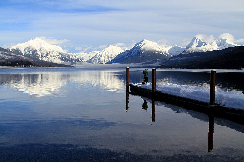 A lone figure stands on a snow-covered dock, overlooking a calm lake with snow-capped mountains reflecting in the water.