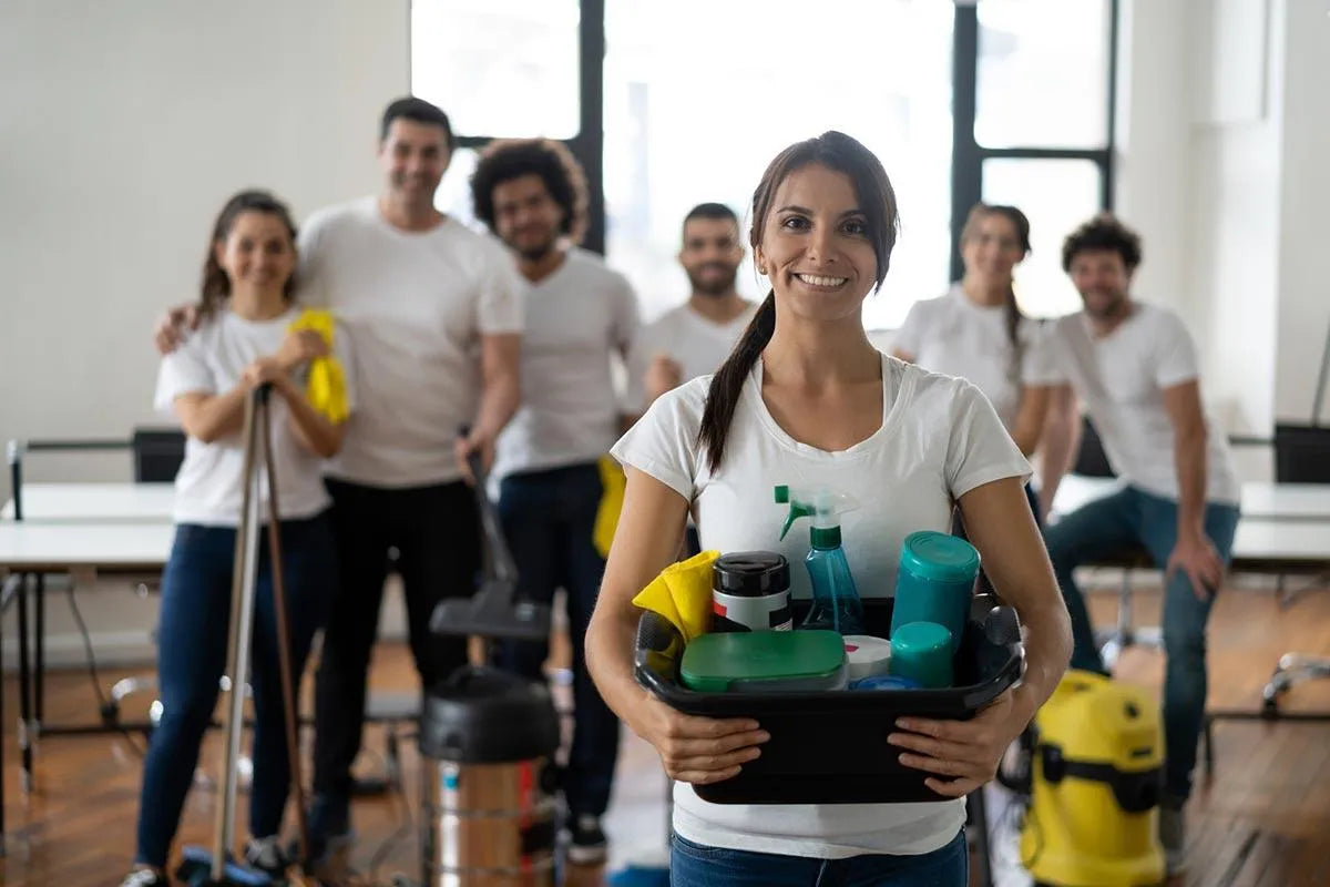  A team of professional cleaners posing in a bright office space, with a smiling woman in the foreground holding a basket of cleaning supplies, and her colleagues equipped with cleaning tools in the background, all wearing casual white t-shirts, representing a friendly and efficient cleaning service.