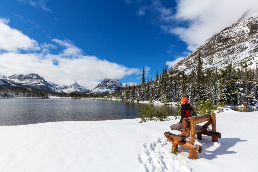 Person sitting on a wooden bench by a snowy lake with mountain views.