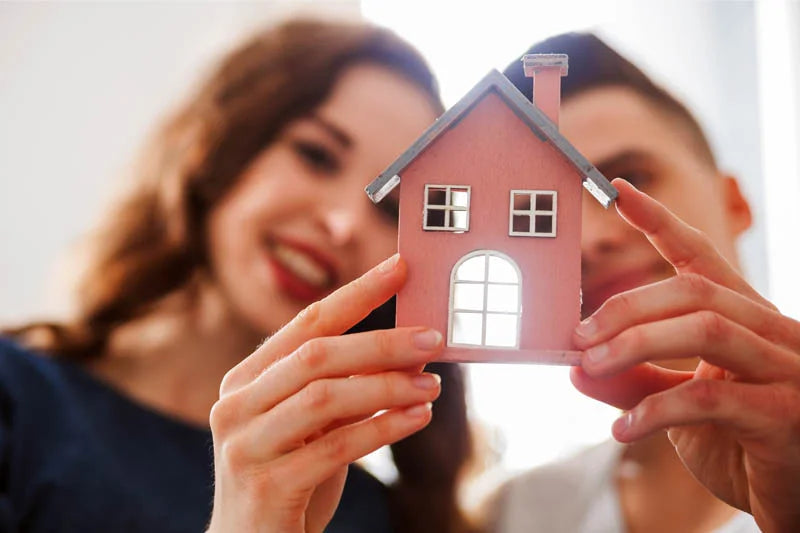 A smiling couple holds a small model house, symbolizing their excitement about buying a new home. The focus is on the house with the couple blurred in the background.