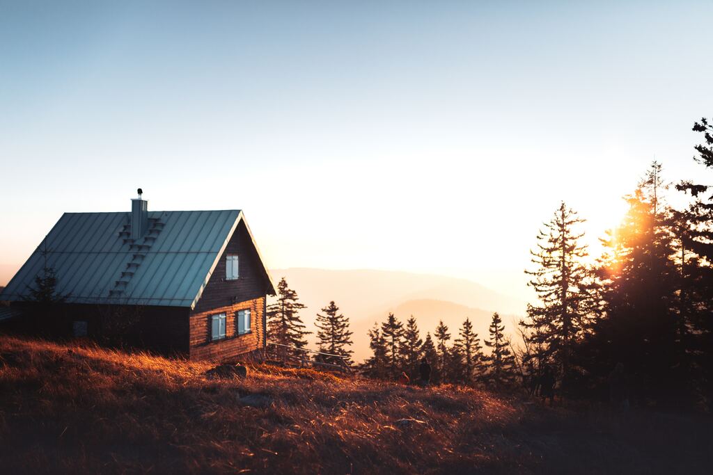 A quaint wooden cabin at sunset, perched atop a hill with golden grasses, flanked by silhouetted pine trees against a soft, glowing sky, offering a serene and picturesque retreat in the wilderness.