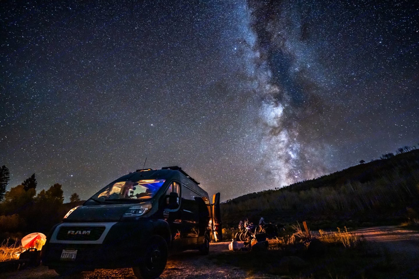  A parked van under the stunning night sky aglow with the Milky Way, adjacent to a campsite with a glowing tent, symbolizing adventure and exploration in the wilderness, with the celestial display highlighting the beauty of stargazing on a clear night.