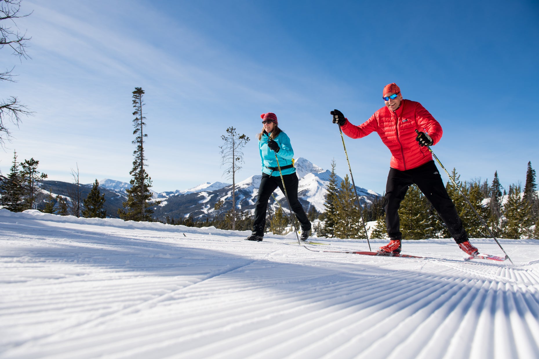 Two people cross-country skiing on a groomed trail with snow-covered mountains in the background.