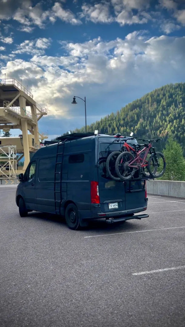  A navy blue van equipped with a rear bike rack carrying two mountain bikes, parked in a lot with a backdrop of lush green hills under a partly cloudy sky, poised for an outdoor cycling adventure.