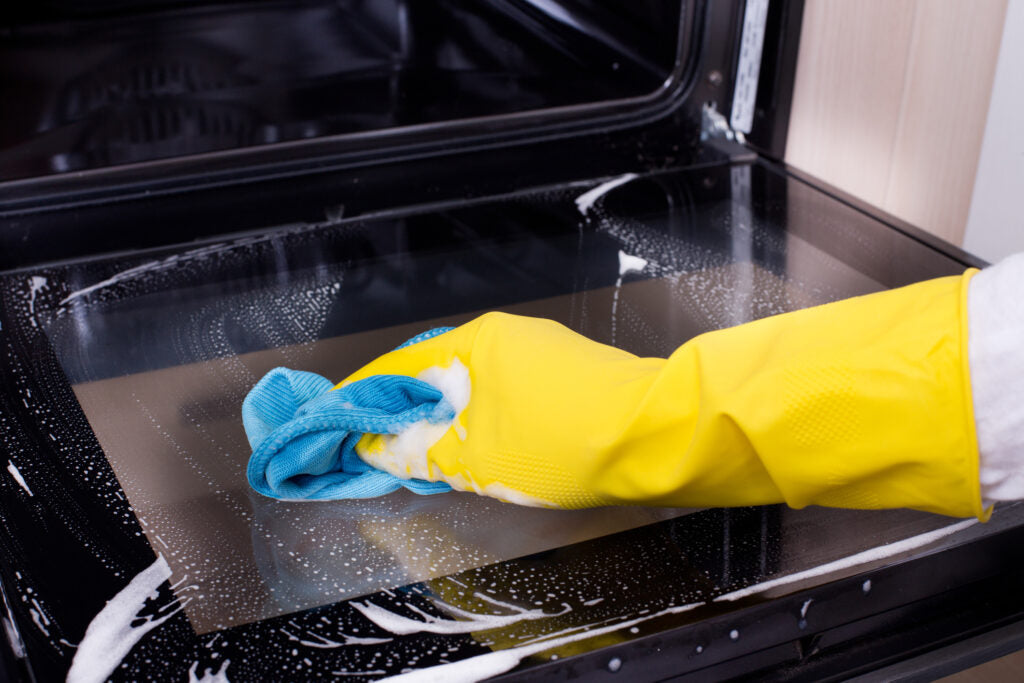 A person wearing a white sleeve and yellow rubber gloves cleaning the inside glass of an oven door with a blue microfiber cloth, depicting a domestic chore and the use of cleaning supplies to maintain kitchen appliances.