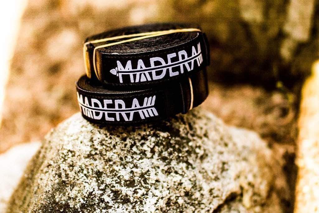 A close-up shot of two rolled black leather belts with the word "MADERA" and an arrow logo printed on them in white, placed on a textured rock in an outdoor setting.