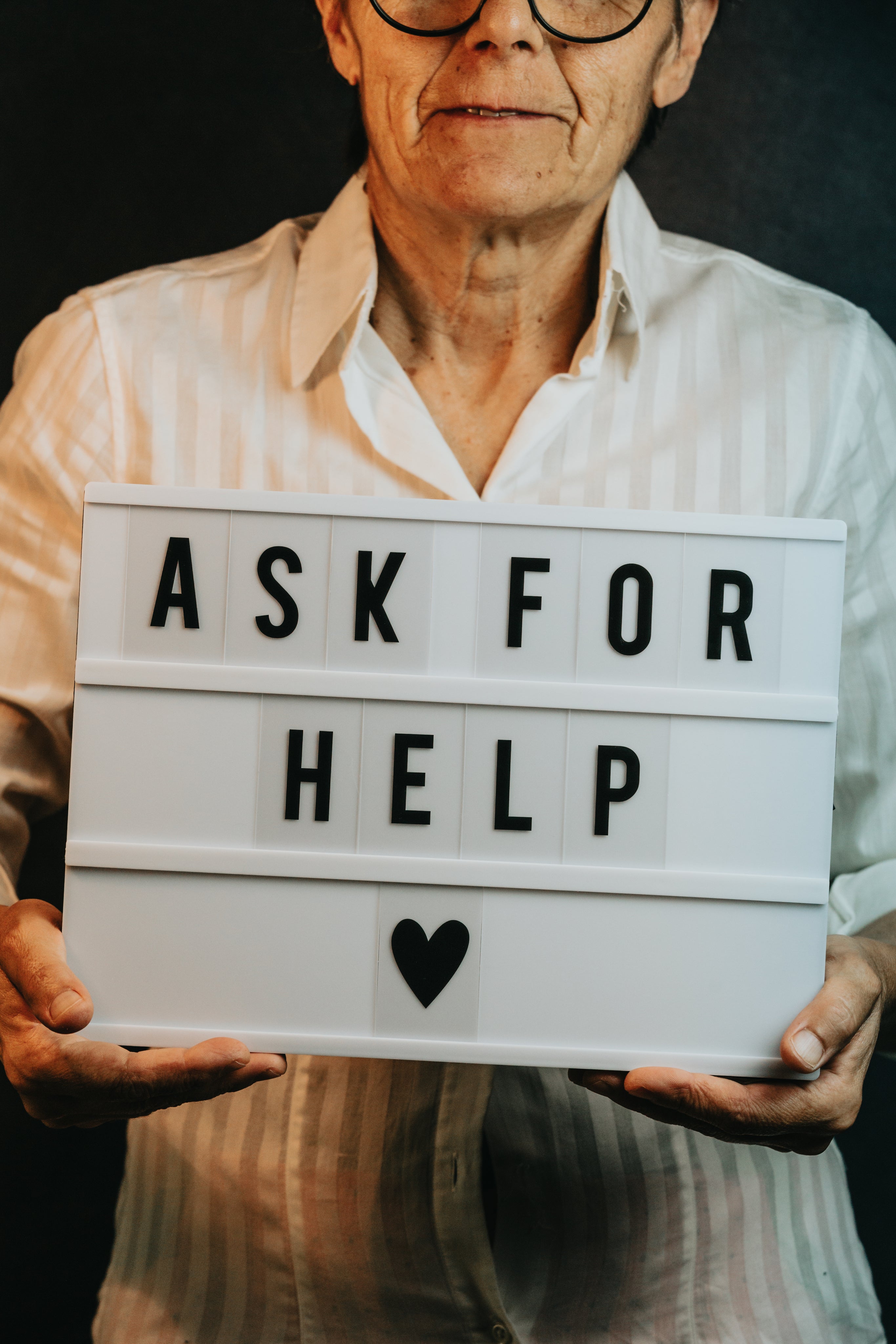 An elder person in a white striped shirt holding a lightbox with the phrase "ASK FOR HELP" displayed in bold letters, emphasizing the importance of seeking support, with a small black heart symbol at the bottom center, against a dark background.