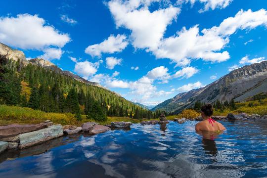 Person relaxing in a hot spring overlooking a scenic mountain valley.