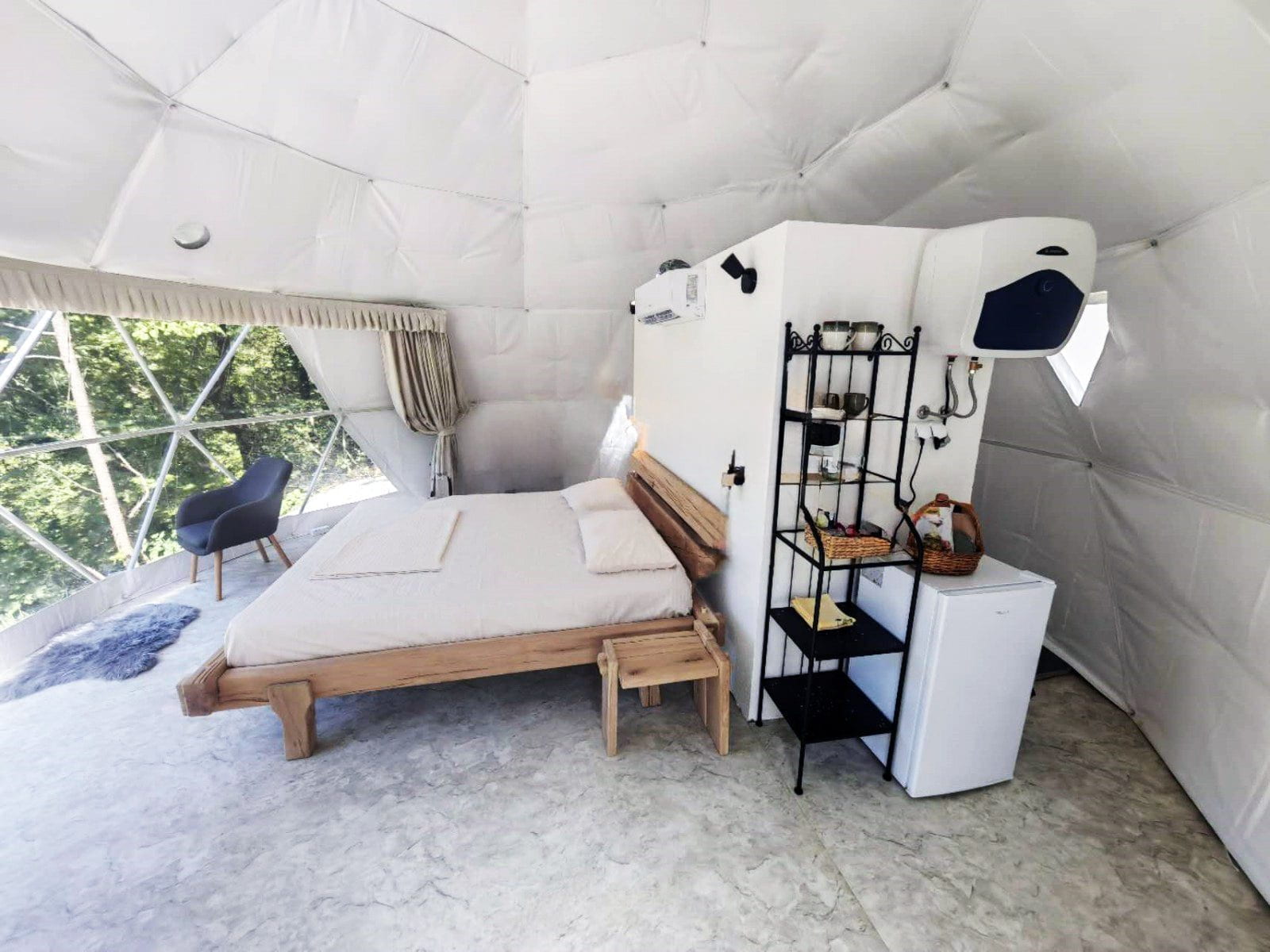 A cozy bedroom inside a geodesic dome tent, featuring a wooden bed with beige linens, a small fridge, and a black metal shelf stocked with essentials. The room has a large window providing a view of the lush greenery outside.