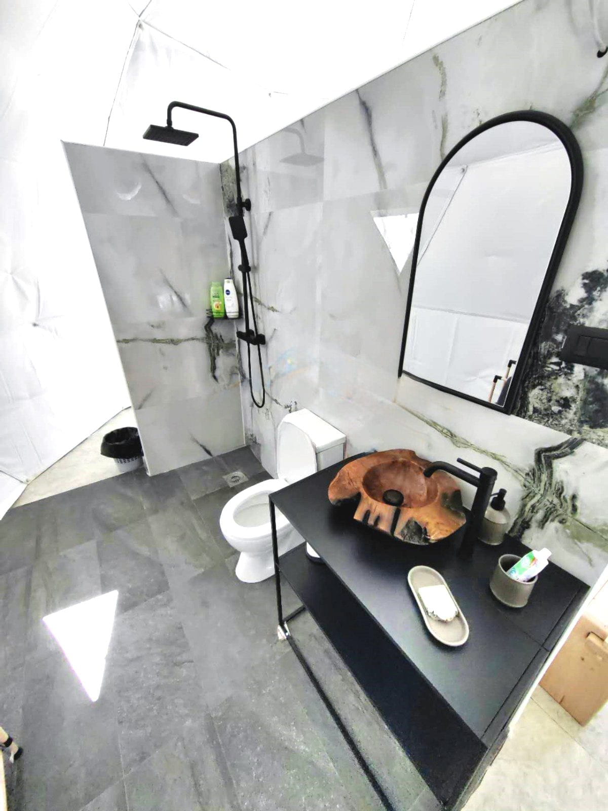 A modern, stylish bathroom with marble walls and floors, featuring a walk-in shower with a black rain showerhead, a sleek black vanity with a wooden basin, and a large oval mirror above it.