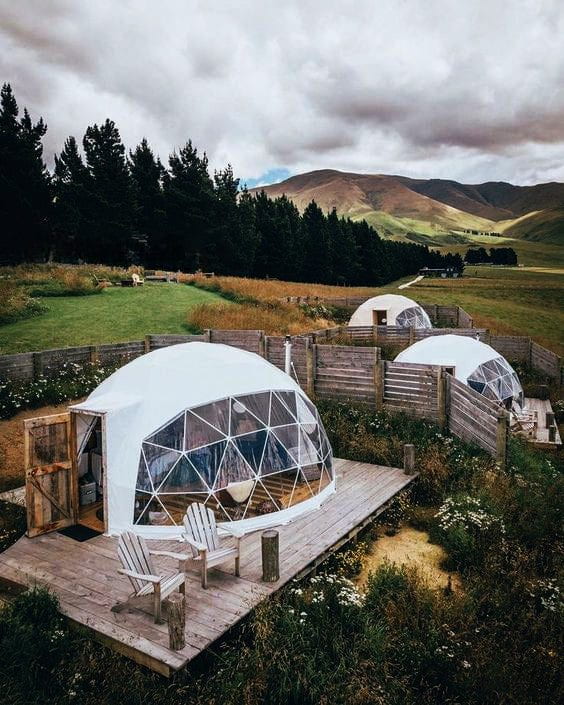 A cluster of white geodesic dome tents situated on wooden decks in a lush, hilly area. The site includes rustic wooden fences, outdoor chairs, and scenic views of mountains and forests, ideal for a tranquil getaway.