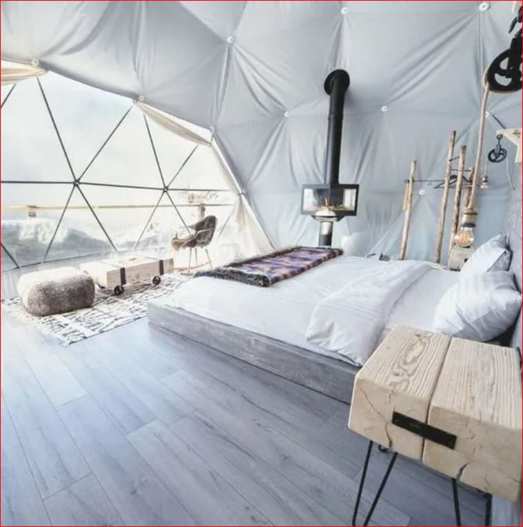 A stylish interior of a geodesic dome tent featuring a modern bed with white linens, a wood-burning stove, and a seating area with a view of a snowy landscape through large windows. The space is decorated with rustic elements, including wooden furniture and hanging lanterns.