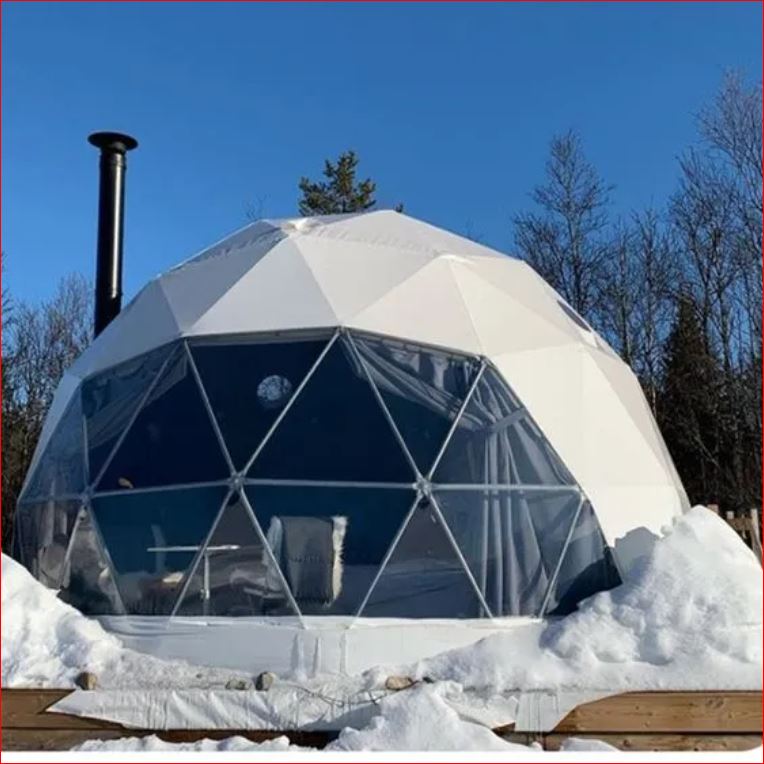 A white geodesic dome tent with large triangular windows, set on a snow-covered wooden deck. The tent has a chimney and is surrounded by a winter landscape with bare trees and a clear blue sky.