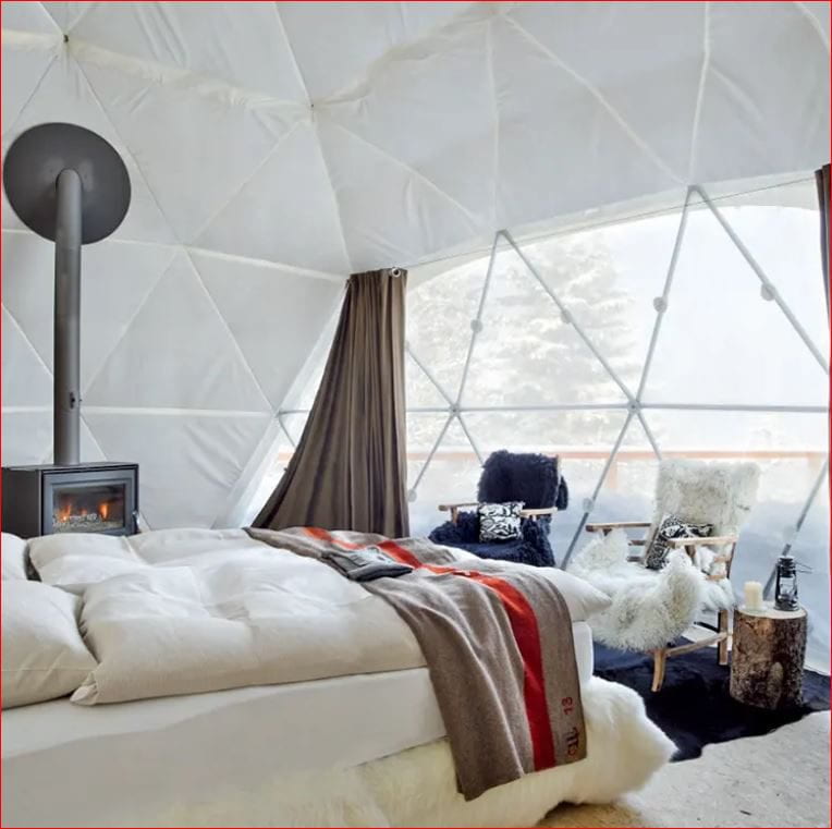 A luxurious interior of a geodesic dome tent featuring a cozy bed with plush white bedding and a colorful blanket, a wood-burning stove for warmth, and a seating area with fur-covered chairs. The large windows offer a picturesque view of the snowy outdoors.