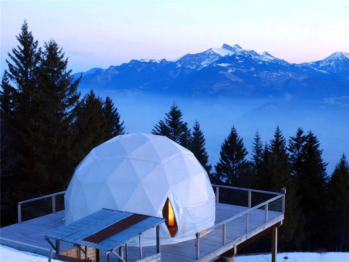 An eco-friendly geodesic dome tent perched on a wooden deck overlooking a breathtaking mountain view. The scene captures the tent amidst a wintery forest with towering pine trees and distant snow-covered peaks.