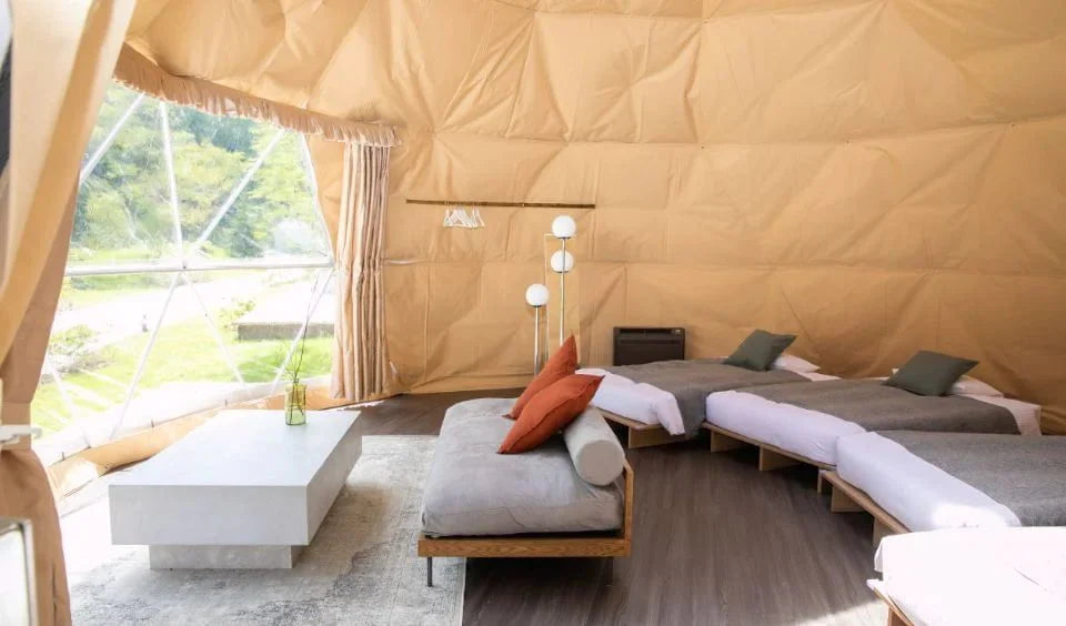 Interior of a spacious geodesic dome tent, featuring multiple single beds with white linens and gray pillows arranged along the wall. 