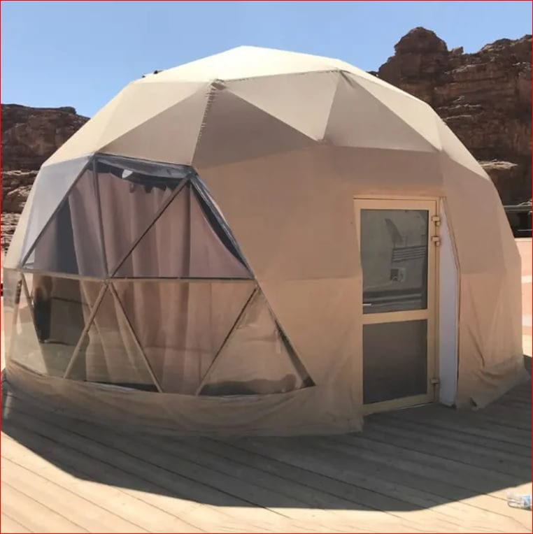 A beige geodesic dome tent with large triangular windows and a door, set on a wooden deck. The tent is situated in a desert-like environment with rocky hills in the background, offering a unique and comfortable glamping experience.